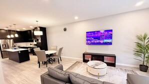 Una sala moderna con un sofá, un comedor con sillas, una cocina con refrigerador y un televisor en la pared.