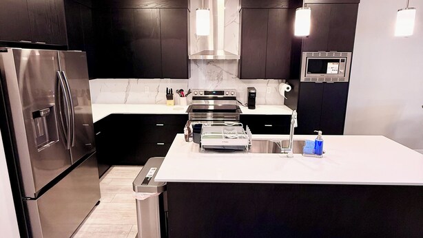 A modern kitchen with dark cabinets, stainless steel appliances, a large island, and pendant lighting. - The Luxe Haven S - Luxury 3BR | 2.5BA Family Home • Stylish • Serene (Calgary)
