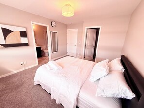 A bedroom with a large bed, a clock on the wall, and a bathroom visible through an open door. - The Luxe Haven S - Luxury 3BR | 2.5BA Family Home • Stylish • Serene (Calgary)