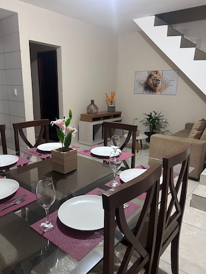 A dining area with a glass table, wooden chairs, and pink placemats. There is a staircase in the background and a framed picture on the wall.