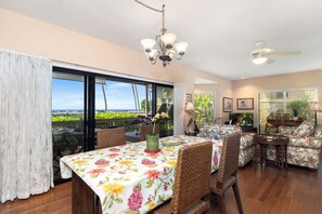 A spacious living room with a large dining table, wicker furniture, and a view of a garden through large windows. - Ocean views at KK2402 Hale Lokahi, a 2-bedroom condo in Keauhou. (Kailua Kona)