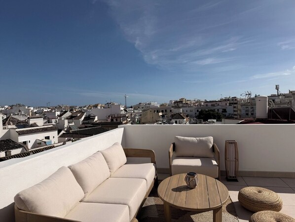Eine Dachterrasse mit einem beigen Sofa, zwei Sesseln, einem runden Holztisch und einer Ziervase. Die Terrasse bietet einen Blick auf eine Stadtlandschaft mit weißen Gebäuden und blauem Himmel.