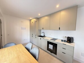 Een moderne keuken met witte kasten, een zwarte spoelbak en een ingebouwde oven. Er staat een houten eettafel met twee stoelen.