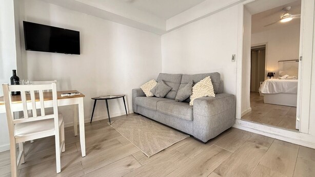 Un salon avec un canapé gris, un fauteuil blanc, une petite table et un téléviseur à écran plat fixé au mur.