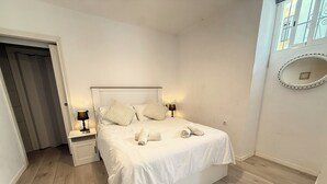 Una cama bien tendida con sábanas y almohadas blancas, una mesita de noche con una lámpara, y un espejo en la pared.