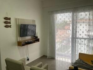 A living room with a flat-screen TV mounted on the wall, a sofa, and a window with a view of buildings outside. - Durei Refuge: Ideal for remote work with fast Wi-Fi! (Paraty)