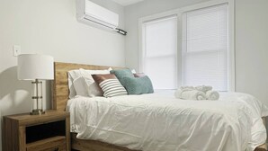 Een slaapkamer met een houten hoofdbord, witte beddengoed, een nachtkastje met een lamp en een wandgemonteerde airconditioner.