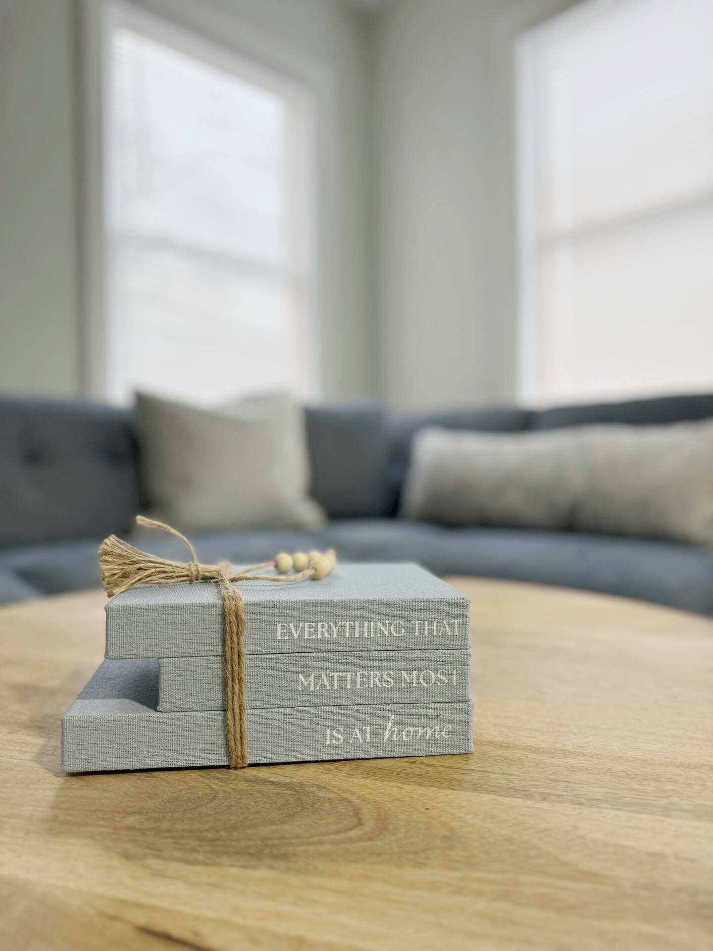 Una pila di scatole di tessuto grigio con la scritta "EVERYTHING THAT MATTERS MOST IS AT HOME" legate con lo spago, posata su un tavolo di legno in una stanza con un divano grigio e una finestra sullo sfondo.