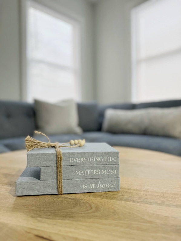 A stack of grey fabric boxes with the text "EVERYTHING THAT MATTERS MOST IS AT HOME" tied with twine, placed on a wooden table in a room with a grey sofa and a window in the background. - Riverside A6 - QUEEN 1BD/1BA Apt near River (Evansville)