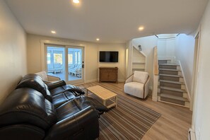 A living room with a black leather sofa, a white armchair, a wooden coffee table, and a TV mounted on the wall. There is a staircase leading to an upper level. - Unique Waterfront Luxury Home - Coastal Entertaining at it's Finest (Scituate)