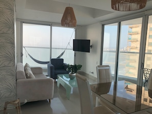 A modern living room with a sofa, armchair, and glass coffee table. A hammock is visible through the large windows, and a TV is mounted on the wall. - Ocean view Appartment 3005 (Right in front of the beach) (Cartagena de Indias)