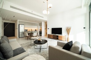 A modern living room with a sofa, coffee table, and TV. There is a kitchen area in the background with a refrigerator and a dining table. - SettlerHomes, Emaar Beachfront Haven with Sea View (Dubai)