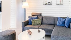 A modern living room with a grey sectional sofa, a white coffee table, and a round ottoman. There are two remote controls on the table, and a lamp is on the left side.