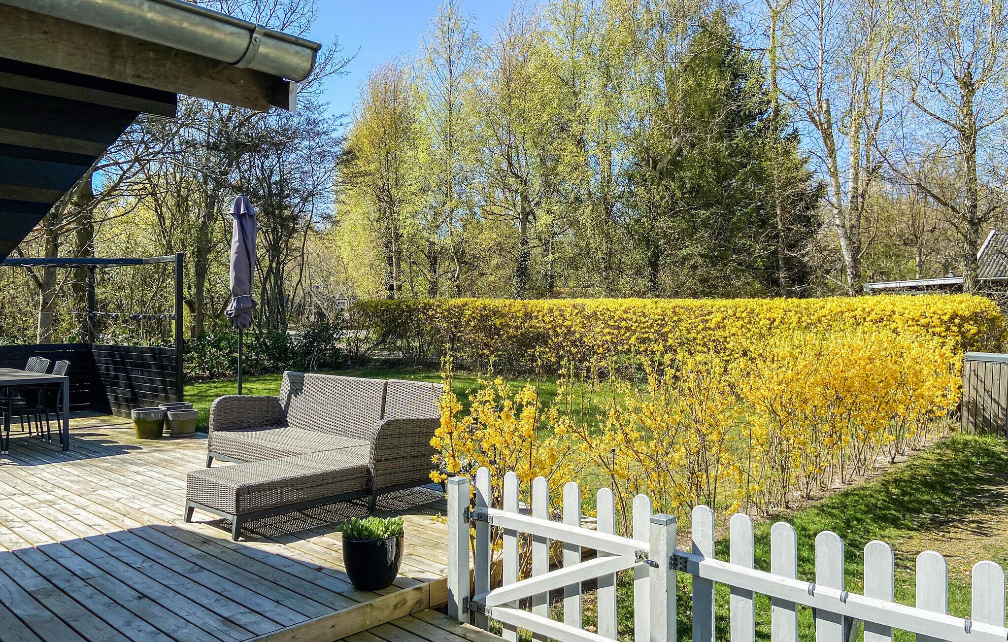 Een houten terras met tuinmeubilair, een wit hek en een goed onderhouden haag van gele bloemen.