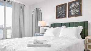 A neatly made bed with white linens and a folded towel, a green upholstered headboard, and two framed floral artworks on the wall.