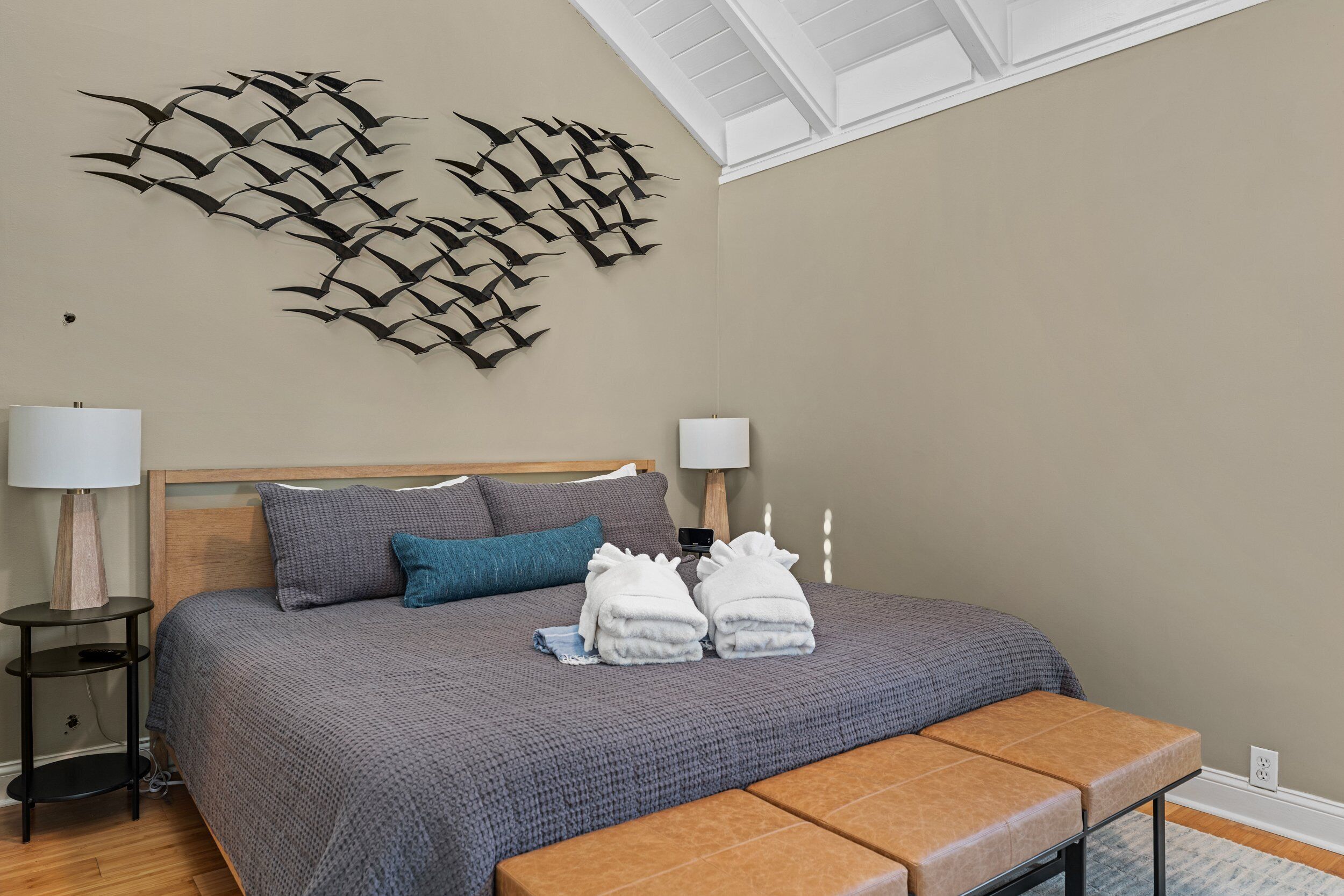 A neatly made bed with a gray blanket, two pillows, and two towels on a wooden bench.