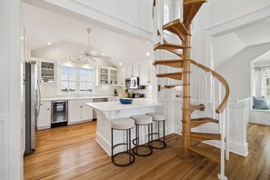Una cocina amplia con armarios blancos, una isla central con taburetes, una escalera espiral de madera y vistas al exterior.