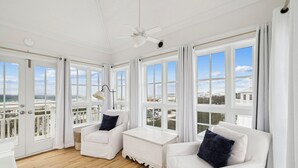 Ein heller, weißer Raum mit großen Fenstern, einem Deckenventilator und zwei weißen Sofas mit dunklen Kissen.