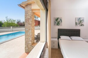 A bedroom with a bed, a nightstand, a window with a view of a swimming pool, and two wall-mounted paintings.