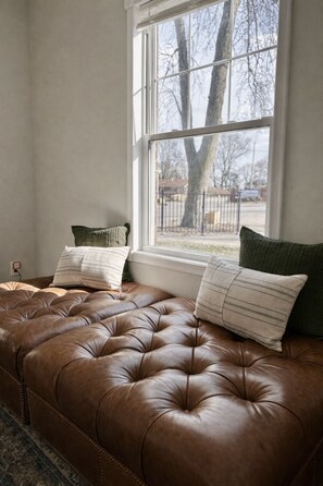 Un banco de cuero marrón con almohadones, una ventana y vistas a árboles y una cerca.
