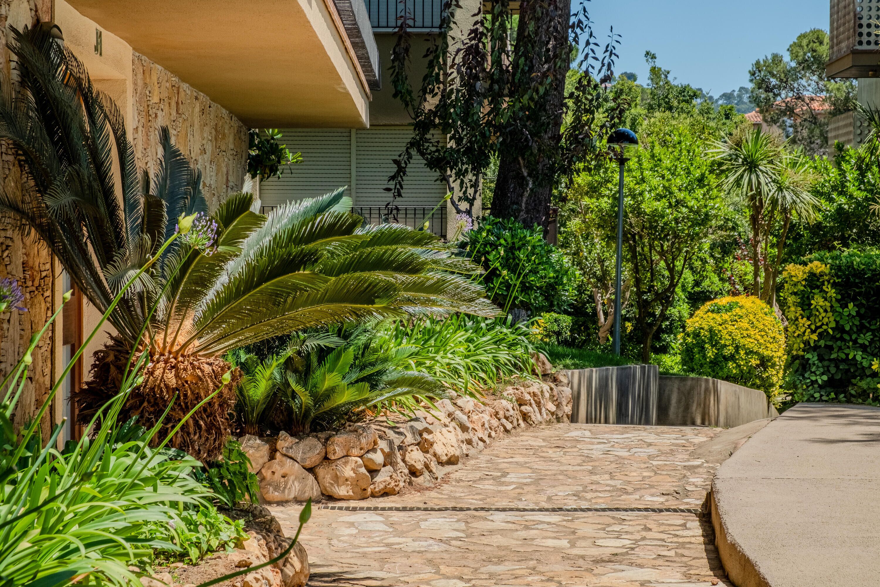 A stone pathway leading to a building with a balcony, surrounded by lush greenery and a clear sky.