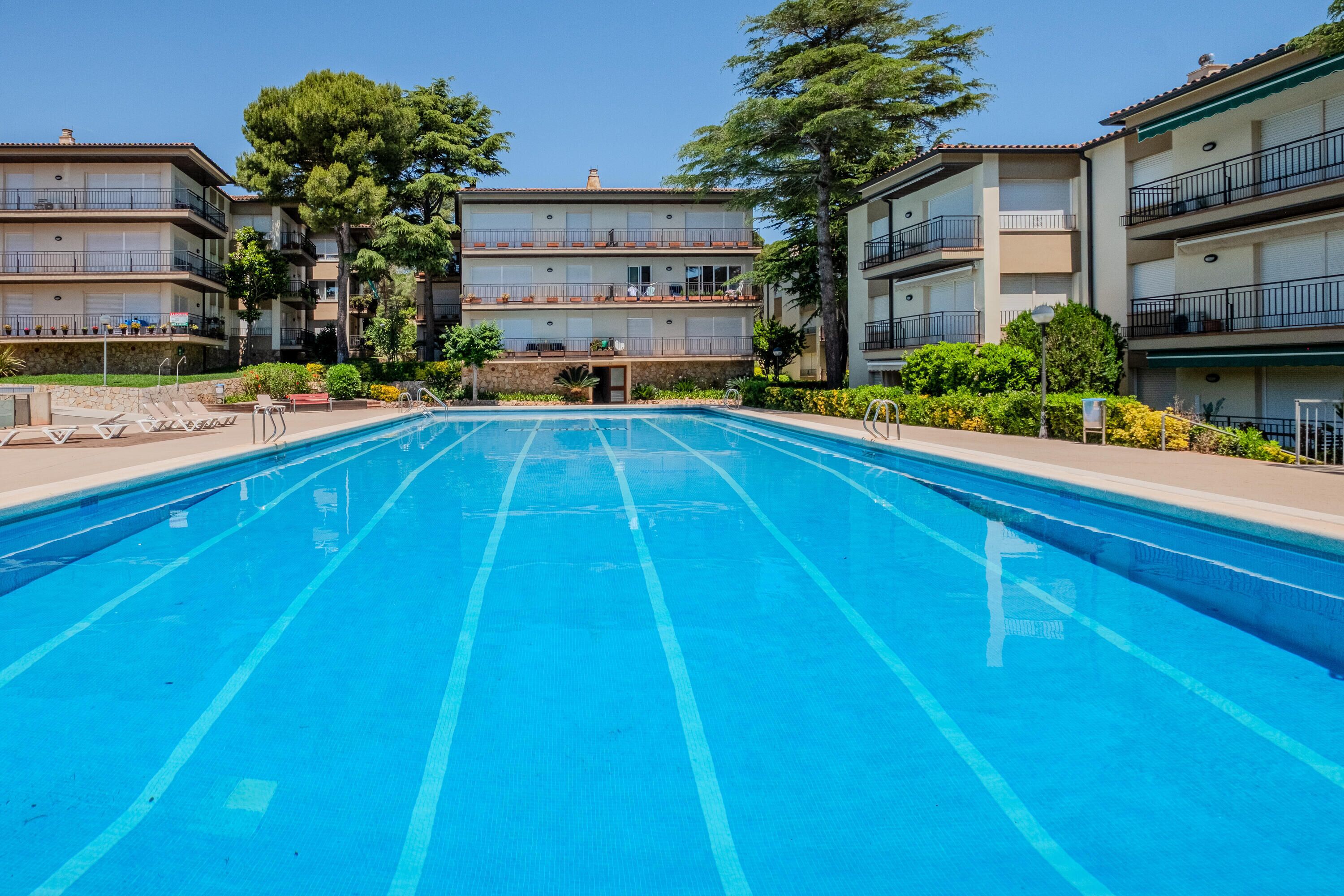 A swimming pool with clear blue water, surrounded by multi-story buildings with balconies, trees, and a stone wall.