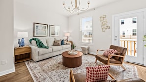 A living room with a sofa, armchair, coffee table, and a chandelier. - Sweetgrass Row at Cane Bay (Summerville)