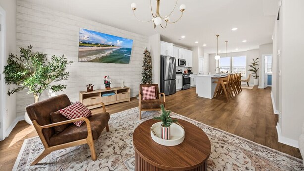 A spacious living area with a fireplace, a dining area with a wooden table, a kitchen with white cabinets, and a large window offering a view. - Sweetgrass Row at Cane Bay (Summerville)