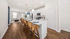 A modern kitchen with white cabinetry, a central island with bar stools, and pendant lighting. - Sweetgrass Row at Cane Bay (Summerville)