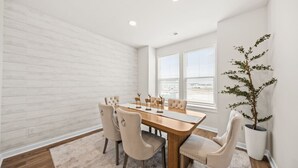 A dining area with a wooden table, beige upholstered chairs, a white brick wall, and a potted plant. - Sweetgrass Row at Cane Bay (Summerville)
