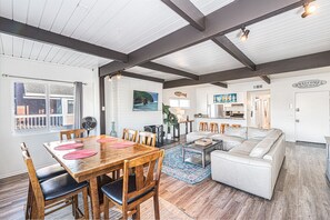 A spacious living area with a wooden dining table, a white sofa, and a kitchen in the background. - 6BR Retreat w/ Expansive Deck, Steps to Beach & Pier! (Newport Beach)