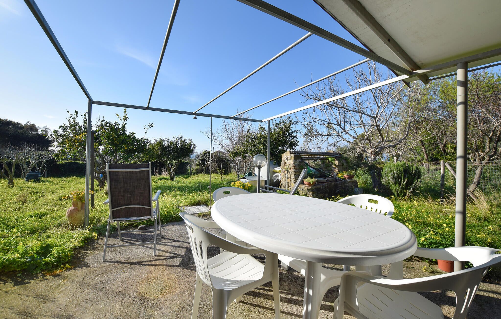 A covered outdoor patio with a round table and chairs, surrounded by greenery and a stone structure.