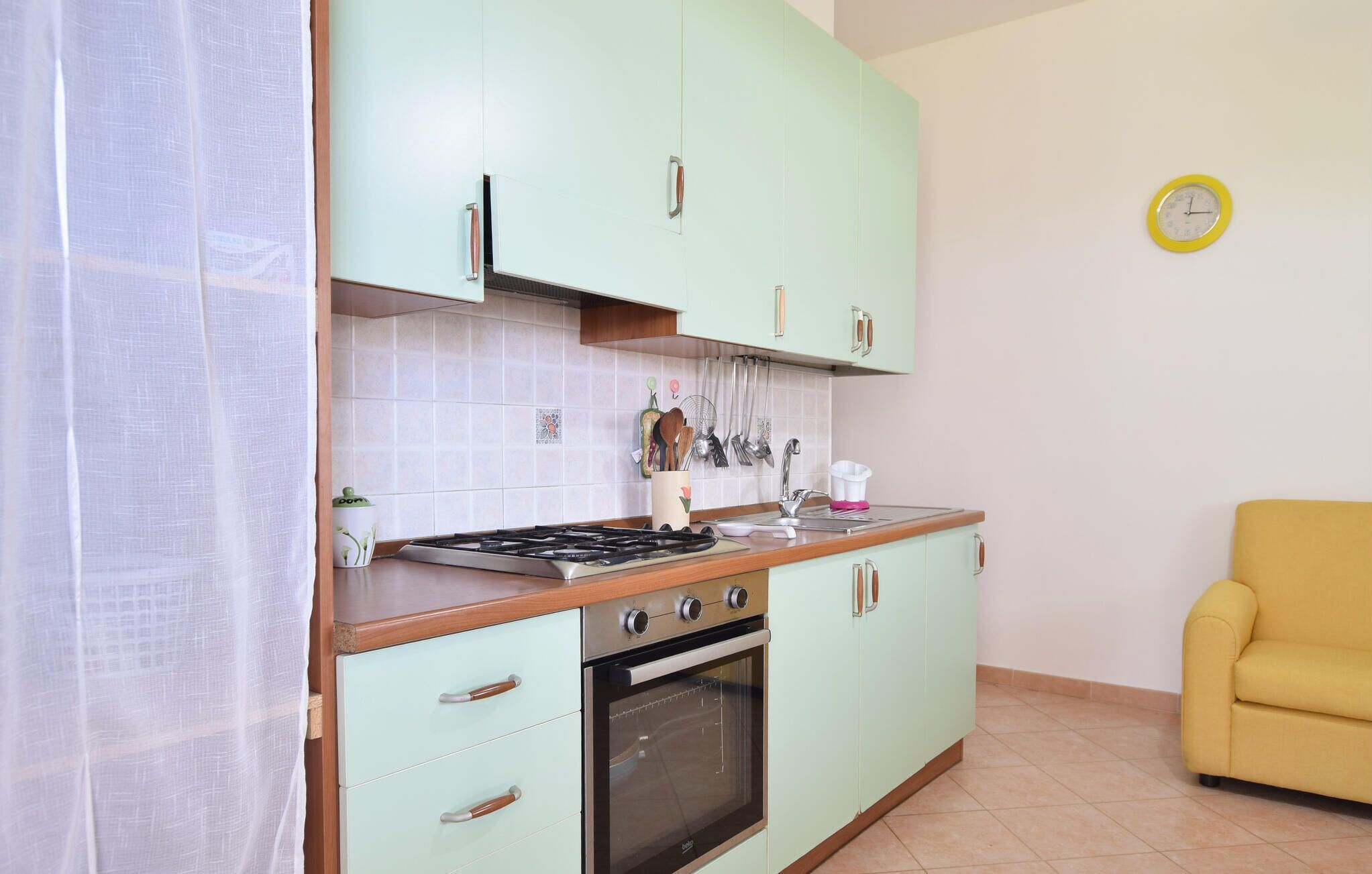 A kitchen with light green cabinets, a stove, an oven, and a yellow chair in the corner.