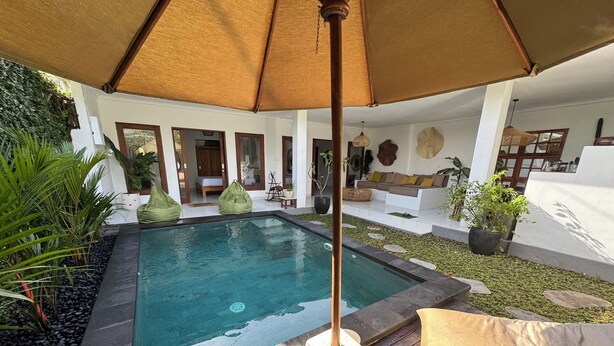 Una piscina con cubierta de madera, rodeada de follaje y un área de estar con cojines.