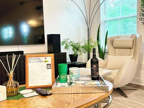 A modern living room with a leather sofa, a flat-screen TV, a speaker, a potted plant, and a tray with a bottle of wine, a remote control, and a framed welcome sign. - New-built Modern home for 2026 Masters rental (Grovetown)