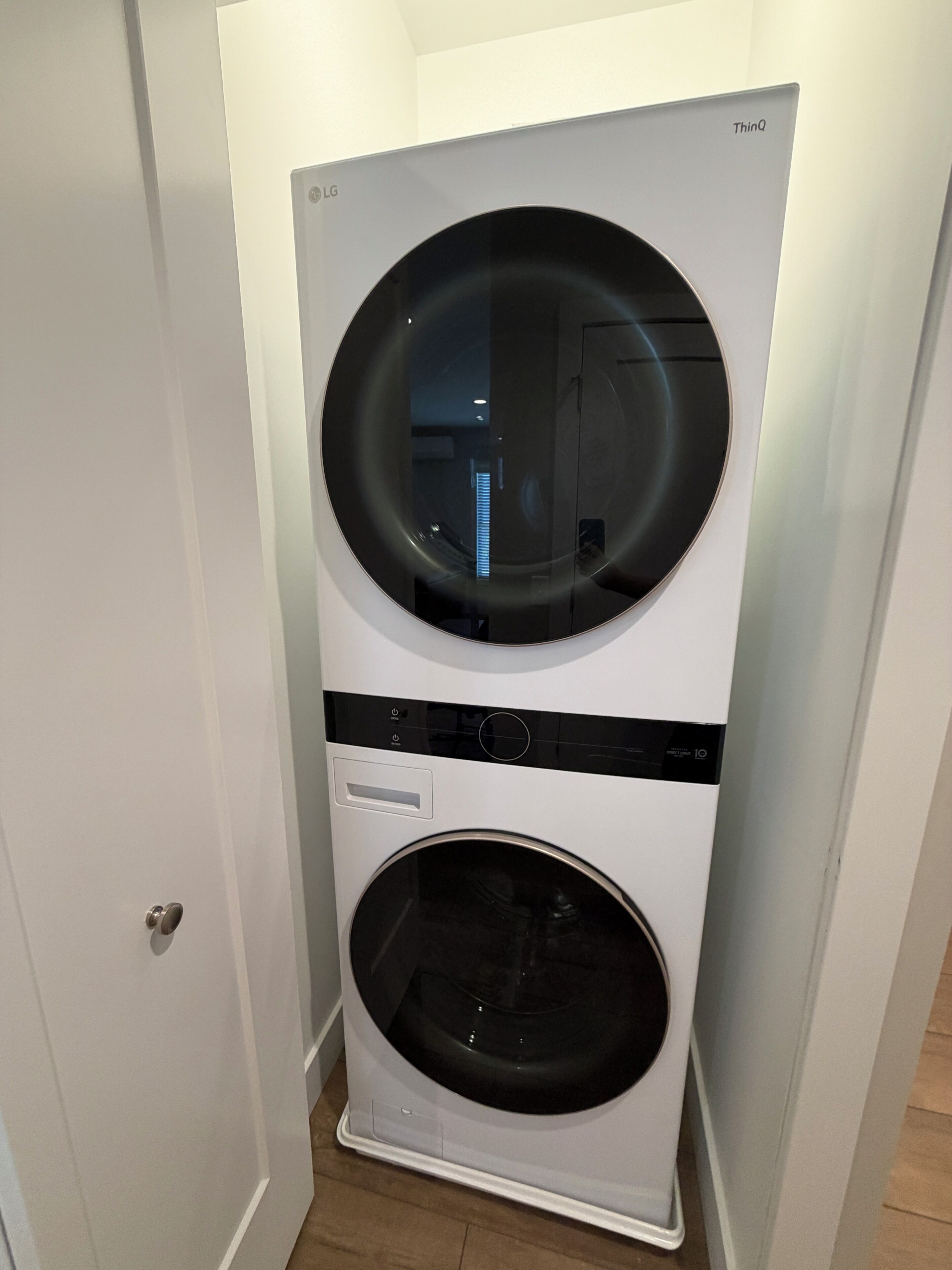 Een witte LG ThinQ wasmachine met een voorlaadontwerp, geplaatst in een hoek van een kamer met een witte deur en houten vloer.