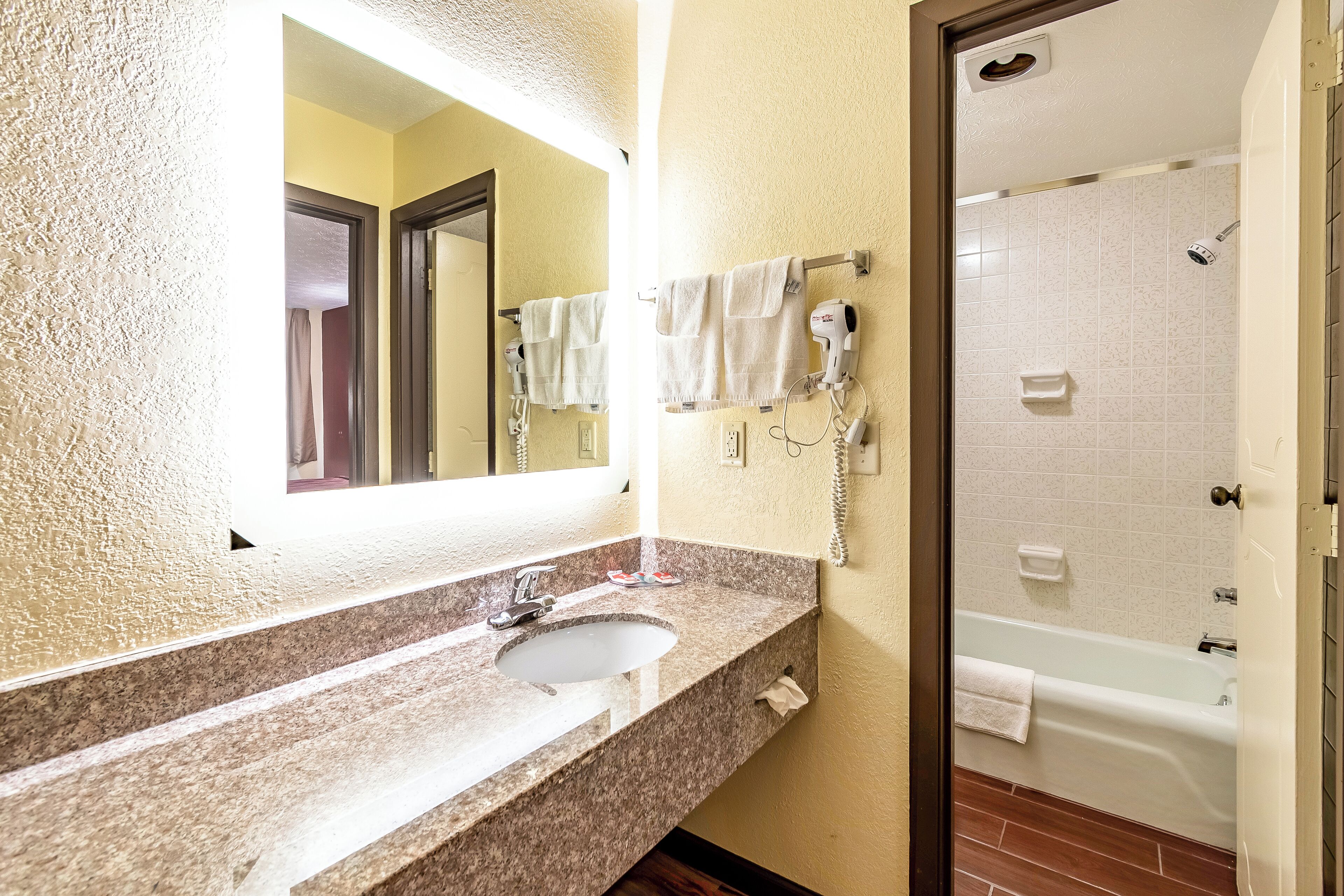 A bathroom with a granite countertop sink, a mirror, a hairdryer, and a shower with a white tub.