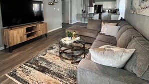 Un salon moderne avec un téléviseur à écran plat, un ventilateur de plafond et une cuisine en arrière-plan.