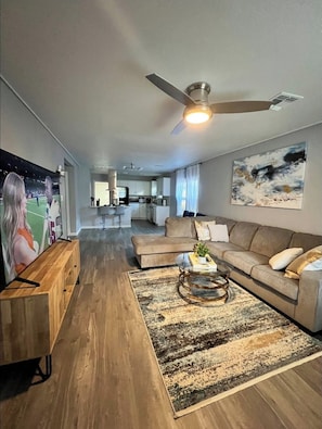 A spacious living room with a large sofa, a coffee table, and a television. There is a kitchen in the background with cabinets and a dining area.