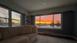 Ein Wohnzimmer mit einem beigefarbenen Sofa, einem gemusterten Teppich und großen Fenstern mit Blick auf einen Sonnenuntergang.