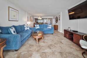 A living room with a blue sofa, a coffee table, a TV, and a dining area in the background. - 2BR 2BA Oceanfront Bliss at Carolina Dunes (Myrtle Beach)