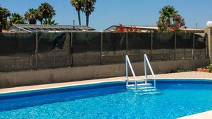 Una piscina amb una escala metàl·lica, envoltada per una tanca, palmeres i un edifici amb teulada solar.