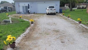 En grusvej fører til et hus med garage, omgivet af krukker med solsikker.