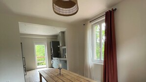 Ein geräumiges Zimmer mit einem Holztisch, einer Bank, einer Hängelampe, einem Fenster mit Vorhängen und Blick ins Freie.
