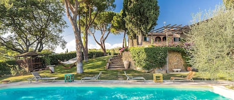 A pool area with a clear blue pool, surrounded by trees and a stone building with a staircase leading up to it.