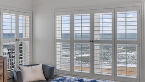 A room with a large window overlooking a beach, a bed with a patterned bedspread, and a chair with a pillow. - NEW listing! Direct Oceanfront 2/2 | Fully Updated | Heated Pool & Garage Parking (Ormond Beach)