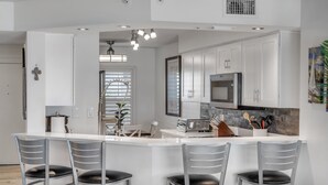 A modern kitchen with white cabinetry, a central island with bar stools, a microwave, and a sink. - NEW listing! Direct Oceanfront 2/2 | Fully Updated | Heated Pool & Garage Parking (Ormond Beach)