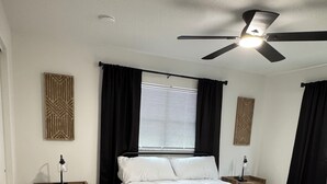 Ein Schlafzimmer mit einem Bett, Deckenventilator, Nachttisch und einem Fenster mit Jalousien.