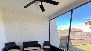Ein Zimmer mit Deckenventilator, drei Korbstühle mit Kissen, einem kleinen Tisch und großen Fenstern mit Blick auf einen Garten mit Holzzaun.
