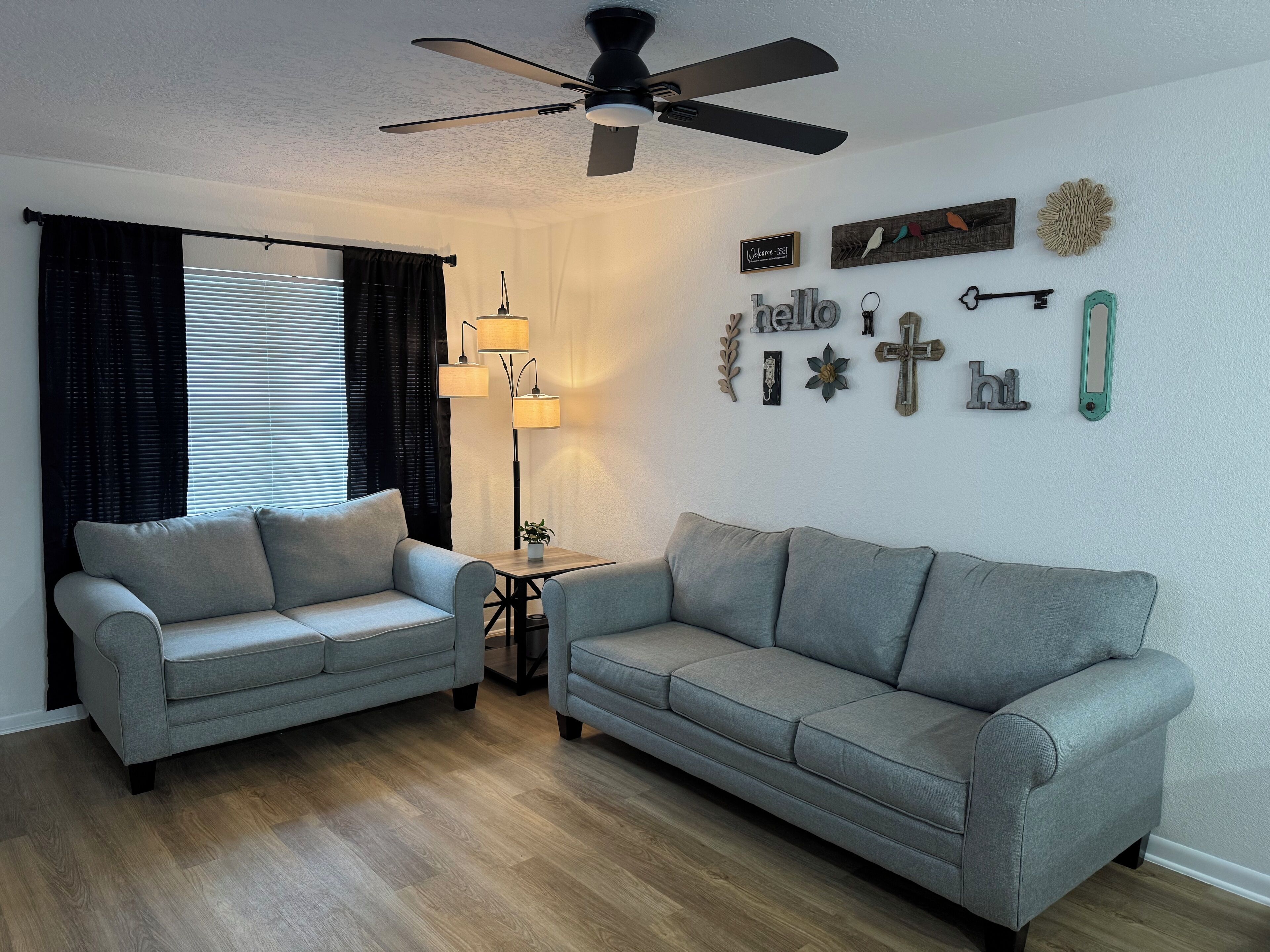 A living room with two grey sofas, a ceiling fan, a floor lamp, and wall decorations including a 'hello' sign and a key.